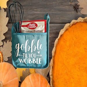May include: Teal oven mitt with the text "gobble til you wobble" in white. A packet of Betty Crocker peanut butter mix is in the pocket of the mitt. A pumpkin pie and pumpkins are in the background.