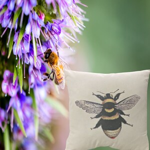 May include: A cushion featuring a detailed illustration of a bee. The bee is black and yellow with detailed wings. The cushion is off-white. A bee is on a purple flower in the background.