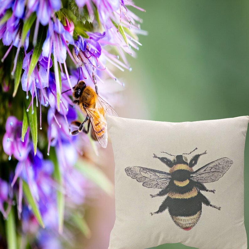 May include: A cushion featuring a detailed illustration of a bee. The bee is black and yellow with detailed wings. The cushion is off-white. A bee is on a purple flower in the background.