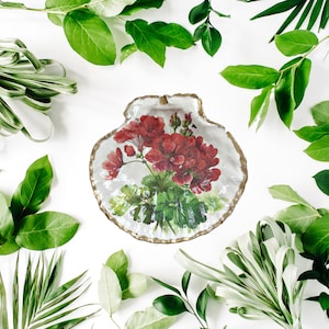 May include: A white ceramic shell-shaped dish with a gold rim, featuring a painted floral design of red geraniums and green leaves.