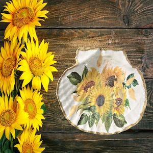 May include: A white ceramic seashell dish with a gold rim, decorated with a painted design of yellow sunflowers and green leaves. The dish is surrounded by a bouquet of yellow sunflowers.