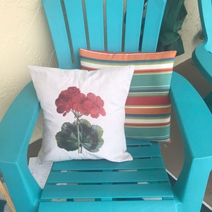 May include: A turquoise plastic adirondack chair with two pillows. The chair has a white pillow with a red geranium print and a striped pillow with orange, red, green, and blue stripes.