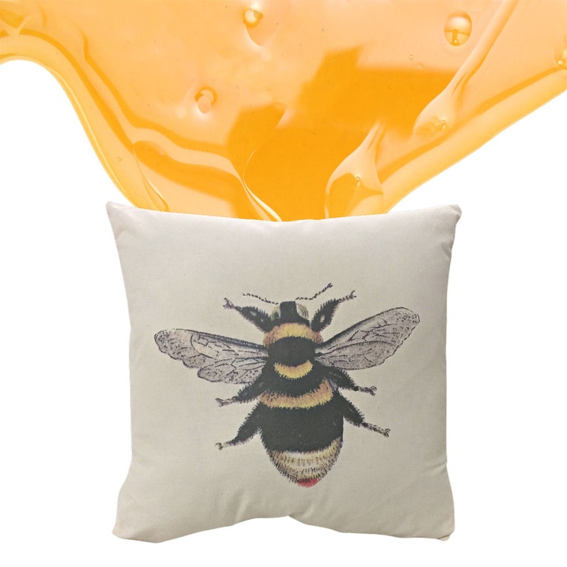 May include: Off-white square cushion with a detailed illustration of a bumblebee. The bee has black and yellow stripes, detailed wings and a red spot. A golden, honey-like substance is above the cushion.