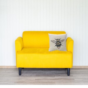 May include: A yellow velvet sofa with black legs and a decorative cushion. The cushion displays a detailed illustration of a bee with black and yellow stripes, wings, and red accents. The background is a white wall.