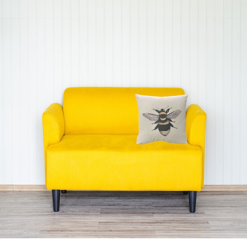 May include: A yellow velvet sofa with black legs and a decorative cushion. The cushion displays a detailed illustration of a bee with black and yellow stripes, wings, and red accents. The background is a white wall.