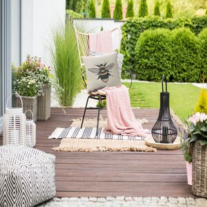 May include: A garden patio scene featuring a chair with a bee-illustrated cushion and a pink throw. The chair is on a wooden deck with a striped rug. A black lantern and potted plants add to the outdoor decor.