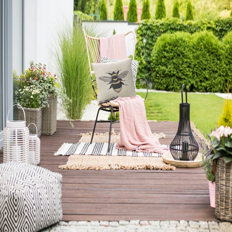 May include: A garden patio scene featuring a chair with a bee-illustrated cushion and a pink throw. The chair is on a wooden deck with a striped rug. A black lantern and potted plants add to the outdoor decor.