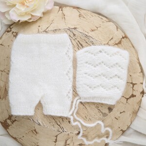 May include: White knitted baby bonnet and matching pants with a delicate lace pattern. The set is displayed on a rustic wooden surface.