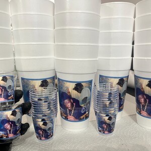 Puede incluir: Una pila de vasos de espuma blancos y vasos de chupito de pl&aacute;stico transparente. Cada vaso y vaso de chupito presenta una etiqueta azul y blanca con una foto de un hombre y el n&uacute;mero 60. Los vasos est&aacute;n apilados.