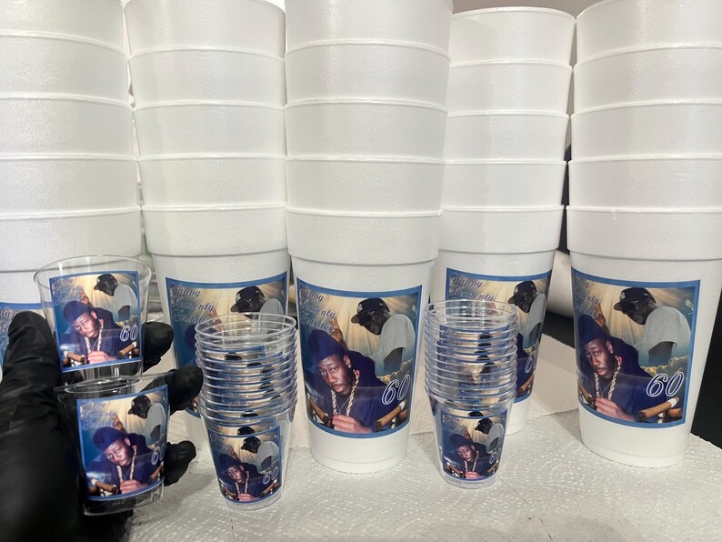 Puede incluir: Una pila de vasos de espuma blancos y vasos de chupito de pl&aacute;stico transparente. Cada vaso y vaso de chupito presenta una etiqueta azul y blanca con una foto de un hombre y el n&uacute;mero 60. Los vasos est&aacute;n apilados.