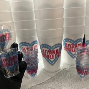 Puede incluir: Una colecci&oacute;n de vasos de espuma blancos y vasos de chupito de pl&aacute;stico transparente. Las tazas y vasos est&aacute;n decorados con un dise&ntilde;o en forma de coraz&oacute;n en azul y rojo, con el nombre "AIYANNA BOULETTE" impreso.