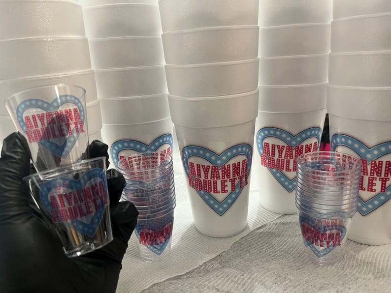 Puede incluir: Una colecci&oacute;n de vasos de espuma blancos y vasos de chupito de pl&aacute;stico transparente. Las tazas y vasos est&aacute;n decorados con un dise&ntilde;o en forma de coraz&oacute;n en azul y rojo, con el nombre "AIYANNA BOULETTE" impreso.