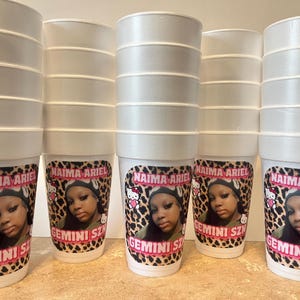 May include: Five white foam cups stacked in columns, each featuring a custom label with a leopard print background, a photograph of a person, a small Hello Kitty character, and the pink text Naima Ariel and Gemini Szn printed on the front.