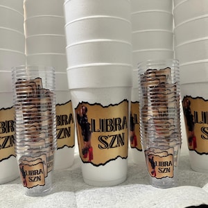 Puede incluir: Una colecci&oacute;n de vasos de espuma blancos y vasos de pl&aacute;stico transparente, cada uno adornado con una etiqueta dorada con el texto "LIBRA SZN" y una imagen de una persona. Los vasos est&aacute;n apilados.
