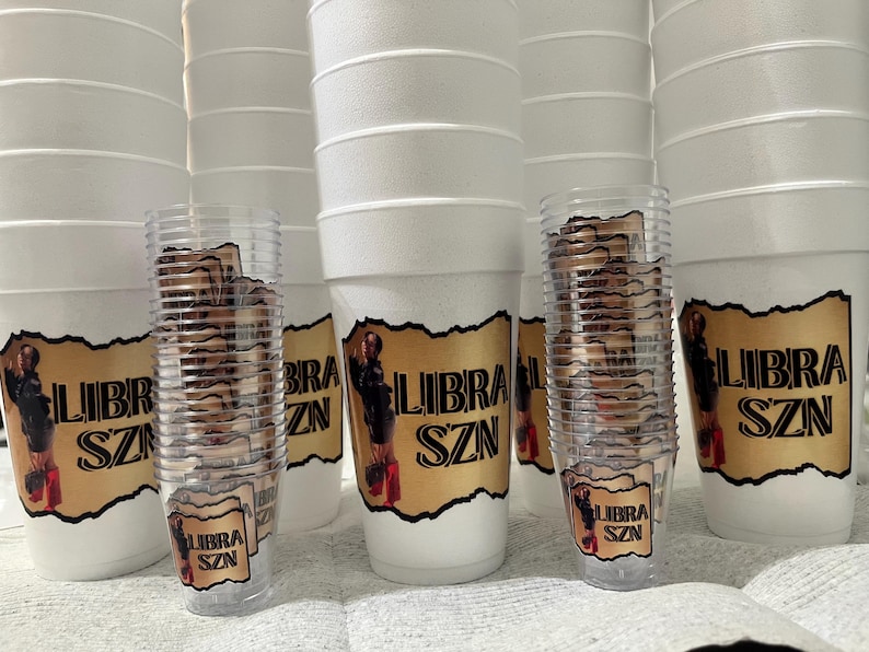 Puede incluir: Una colecci&oacute;n de vasos de espuma blancos y vasos de pl&aacute;stico transparente, cada uno adornado con una etiqueta dorada con el texto "LIBRA SZN" y una imagen de una persona. Los vasos est&aacute;n apilados.