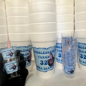 Puede incluir: Una colecci&oacute;n de vasos de espuma blancos y vasos de chupito de pl&aacute;stico transparente. Las tazas y los vasos est&aacute;n decorados con una etiqueta azul y blanca que dice "Bubblessaid Takes 32 Libra Season". Imagen de cerca.