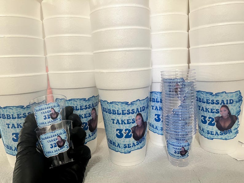 Puede incluir: Una colecci&oacute;n de vasos de espuma blancos y vasos de chupito de pl&aacute;stico transparente. Las tazas y los vasos est&aacute;n decorados con una etiqueta azul y blanca que dice "Bubblessaid Takes 32 Libra Season". Imagen de cerca.