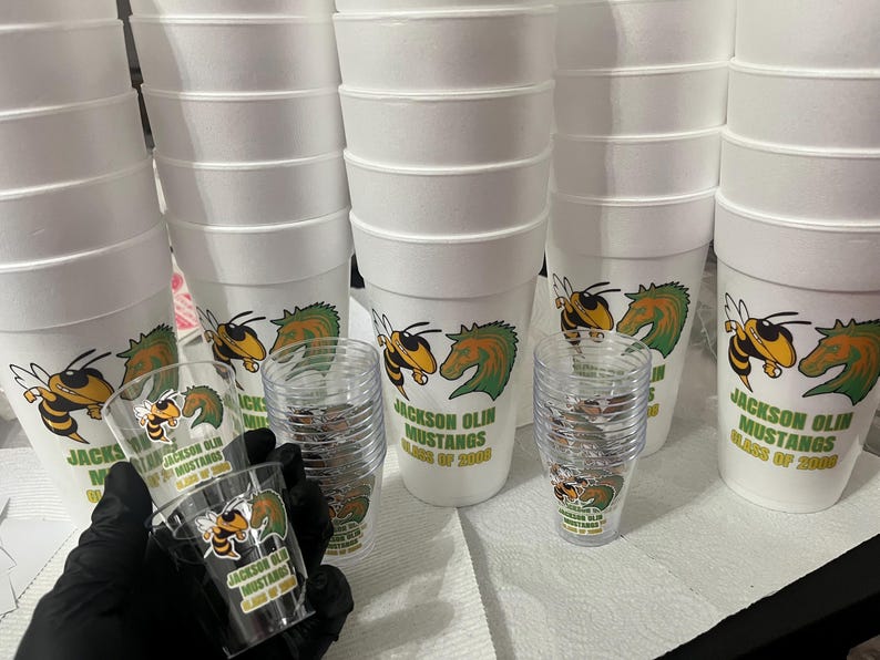 Puede incluir: Vasos de espuma blancos y vasos de chupito de pl&aacute;stico transparente con un logotipo de Jackson Olin Mustangs con una abeja y una cabeza de caballo en verde y amarillo. El texto dice "Jackson Olin Mustangs Class of 2000".