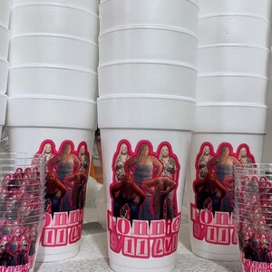 Puede incluir: Vasos de espuma blancos y vasos de pl&aacute;stico transparente con un gr&aacute;fico rosa y rojo. El gr&aacute;fico presenta un grupo de mujeres y el texto "RONNIE VILLA". Los vasos est&aacute;n apilados y dispuestos en grupo.