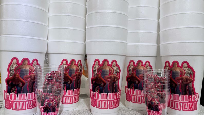 Puede incluir: Vasos de espuma blancos y vasos de pl&aacute;stico transparente con un gr&aacute;fico rosa y rojo. El gr&aacute;fico presenta un grupo de mujeres y el texto "RONNIE VILLA". Los vasos est&aacute;n apilados y dispuestos en grupo.