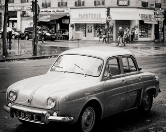 Op de afbeelding: Een zwart-witfoto van een vintage auto geparkeerd op een straat in Parijs, Frankrijk. De auto is een Renault Dauphine, een kleine vierdeurs sedan. De auto heeft een kentekenplaat met de tekst "AS 991 TL". De auto staat geparkeerd voor een gebouw met een bord met de tekst "Papeterie".