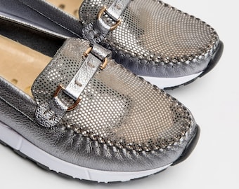 May include: A pair of silver leather loafers with a perforated design and a gold buckle. The loafers have a white rubber sole.