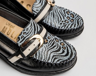 May include: Black leather loafers with a zebra print and gold buckle. The loafers have a white stripe across the front and the text "Unique Molas" is printed on the inside.