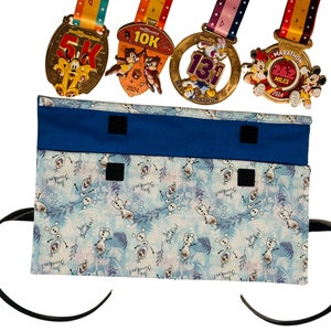Puede incluir: Una bolsa de tela azul con un forro de tela blanco con un patr&oacute;n repetido de Olaf de Frozen. La bolsa tiene dos cierres de velcro negros y una correa negra. Cuatro medallas de oro con cintas de colores se muestran encima de la bolsa.