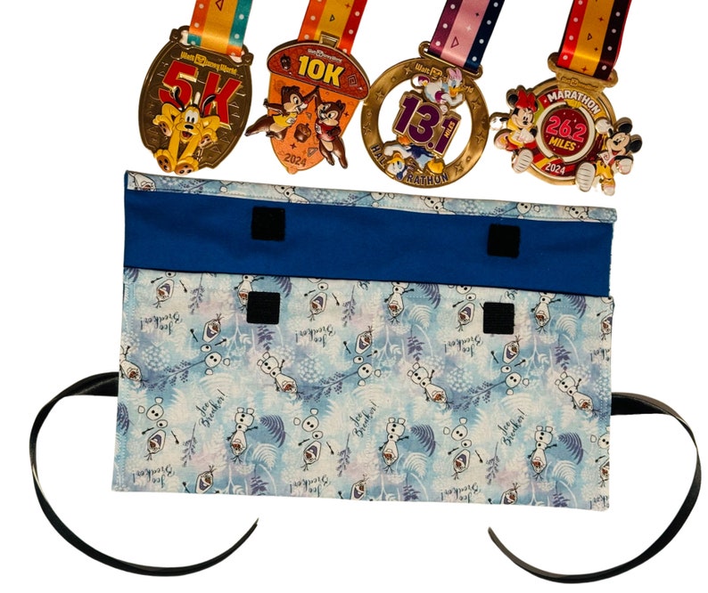 Puede incluir: Una bolsa de tela azul con un forro de tela blanco con un patr&oacute;n repetido de Olaf de Frozen. La bolsa tiene dos cierres de velcro negros y una correa negra. Cuatro medallas de oro con cintas de colores se muestran encima de la bolsa.