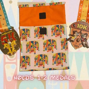 May include: A bright orange fabric pouch with a white and colorful print of a castle. The pouch has two velcro closures and is designed to hold 1-2 medals. The text "HOLDS 1-2 MEDALS" is printed below the pouch.