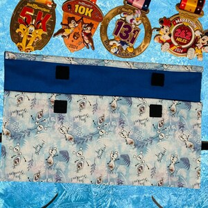 Puede incluir: Una bolsa de tela azul con cierres de velcro, con un estampado de Frozen de Olaf con el texto "You're a good snowman, Olaf!" La bolsa est&aacute; dise&ntilde;ada para guardar y organizar medallas.