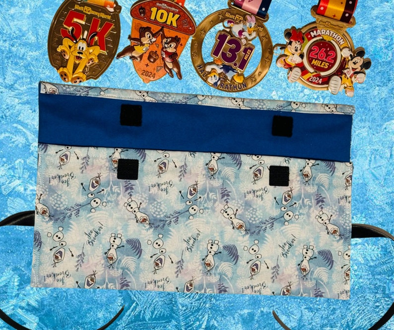 Puede incluir: Una bolsa de tela azul con cierres de velcro, con un estampado de Frozen de Olaf con el texto "You're a good snowman, Olaf!" La bolsa est&aacute; dise&ntilde;ada para guardar y organizar medallas.