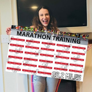May include: A woman holds up a red and white marathon training chart with 26.2 miles printed at the bottom. The chart is divided into 28 weeks and includes daily training plans.