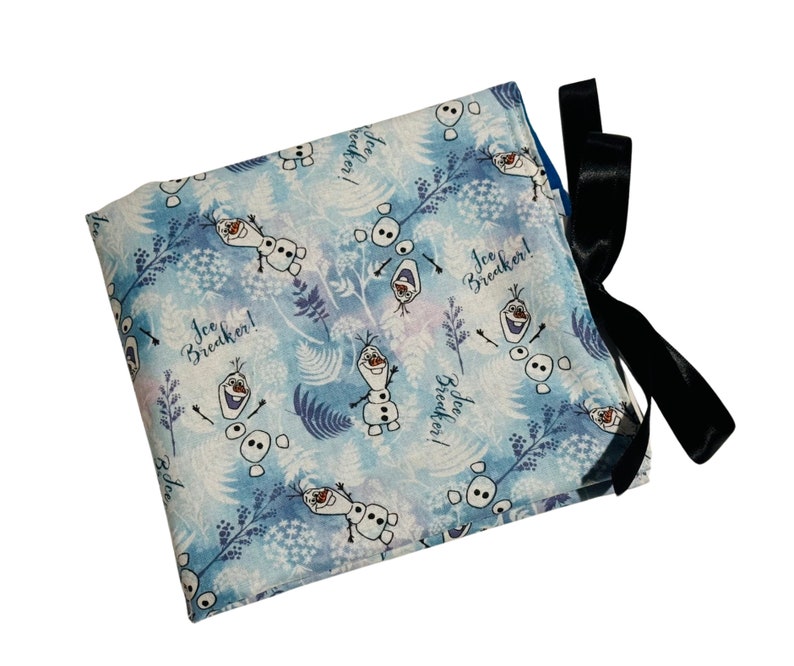 Puede incluir: Una bolsa de tela azul y blanca con un lazo negro. La bolsa presenta un patr&oacute;n repetido de Olaf de la pel&iacute;cula Frozen, con el texto "Ice Breaker!" repetido en toda la superficie.