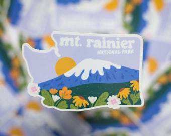 May include: A sticker featuring a stylized illustration of Mount Rainier National Park in Washington state. The sticker is shaped like the state of Washington and features a purple sky, a white mountain peak, a yellow sun, and green trees and flowers at the base of the mountain. The text "mt. rainier NATIONAL PARK" is printed in black at the top of the sticker.