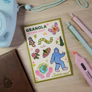 May include: A light blue Instax Mini 9 camera, brown leather journal, sticker sheet with illustrations, pens, and a pink highlighter on a wooden surface. The sticker sheet says "Granola" and features a Bigfoot illustration.