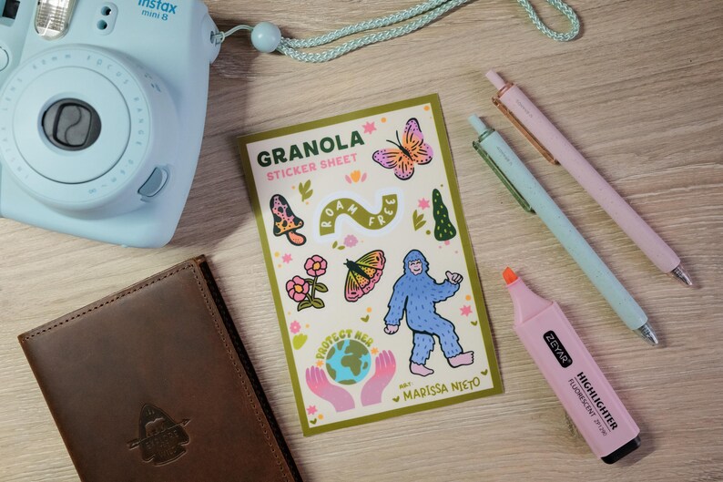 May include: A light blue Instax Mini 9 camera, brown leather journal, sticker sheet with illustrations, pens, and a pink highlighter on a wooden surface. The sticker sheet says "Granola" and features a Bigfoot illustration.