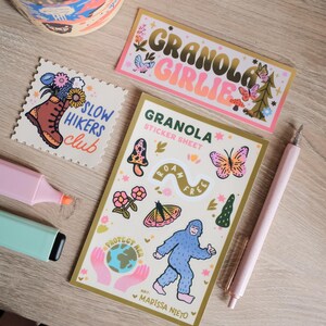 May include: A collection of stickers and stationery items, including a sticker sheet with a Bigfoot illustration, a sticker that says "Granola Girlie", and a sticker with a hiking boot and flowers. Also included are two highlighters and a pink pen.