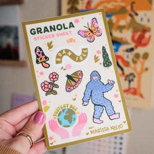 May include: A sticker sheet with a gold border featuring the word "GRANOLA" and the text "STICKER SHEET". The sheet includes illustrations of a butterfly, mushroom, flowers, a bigfoot, and the text "ROAM FREE" and "PROTECT HER".