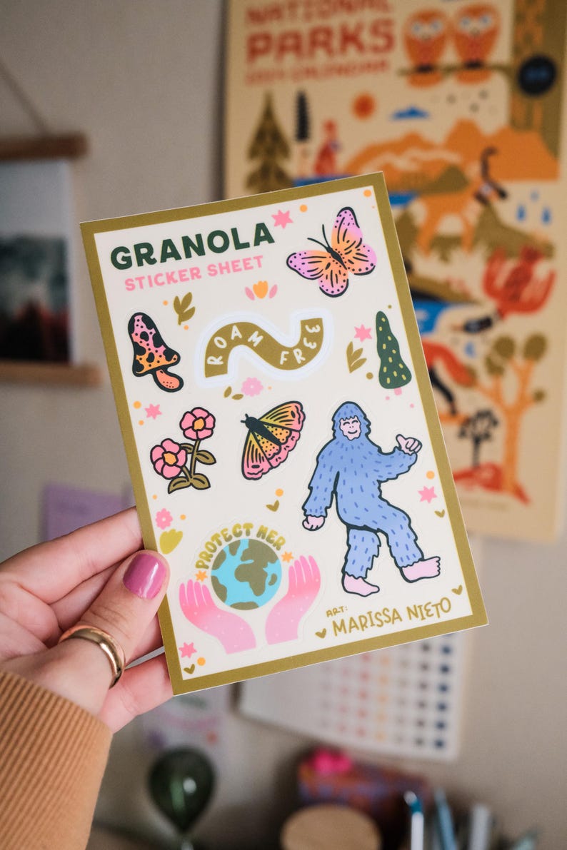 May include: A sticker sheet with a gold border featuring the word "GRANOLA" and the text "STICKER SHEET". The sheet includes illustrations of a butterfly, mushroom, flowers, a bigfoot, and the text "ROAM FREE" and "PROTECT HER".