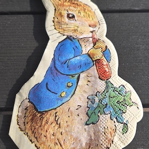 May include: A paper napkin featuring a cartoon illustration of Peter Rabbit. The rabbit is wearing a blue jacket and holding a carrot. The napkin has a textured surface and is set against a dark background.