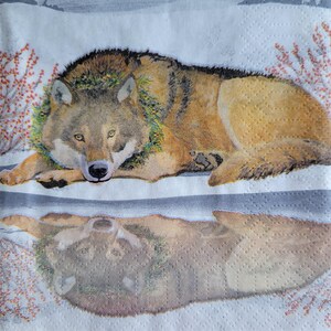May include: A decorative paper napkin featuring a detailed illustration of a wolf lying down. The wolf has brown and tan fur, a green wreath around its neck, and is set against a snowy background with red berry branches.