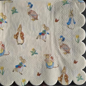 May include: A square, off-white paper napkin with a scalloped edge. The napkin features illustrations of rabbits, ducks, mice, and flowers in pastel colors. The design is reminiscent of a children's storybook.