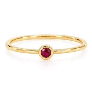Könnte beinhalten: Ein dünner, polierter Gelbgoldring mit einem einzelnen, kleinen, runden und facettierten tiefroten Rubin, der in einer glatten, kreisförmigen Zargenfassung in der Mitte des Rings sitzt und ein schlichtes, elegantes Schmuckstück bildet.