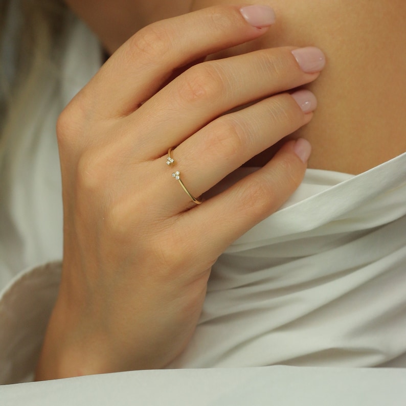 May include: A gold ring with two small diamonds on the band. The ring is open and sits on the finger.