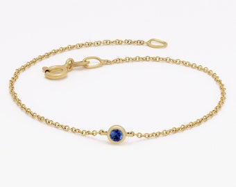 May include: A delicate gold chain bracelet featuring a single round, bezel-set blue gemstone at the center. The bracelet includes a circular spring ring clasp and a small jump ring at the end of the fine, polished gold link chain.