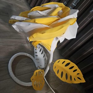 Peut inclure: Une grande rose en papier faite à la main, jaune et blanche. La fleur a une tige et des feuilles blanches, avec une feuille jaune avec des détails découpés. La rose est éclairée par un cordon blanc et un interrupteur.