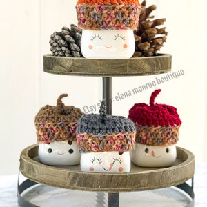 May include: Three white ceramic mugs with smiling faces, each wearing a different colored crocheted acorn hat. The hats are brown, gray, and red. The mugs are displayed on a two-tiered wooden stand.