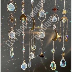 Puede incluir: Atrapasoles dorados con detalles de cristal transparente e iridiscente. Cada atrapasoles presenta una combinación de esferas de cristal, lágrimas y formas geométricas, junto con amuletos de temática celestial como lunas y estrellas. El nombre de la tienda Etsy es visible.