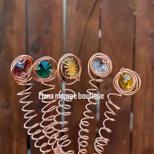 May include: Close-up of five copper hair spirals, each with a different colored gemstone or decorative element at the top. The gemstones are red, green, clear, and yellow. The text "Elena monroe boutique" is visible.